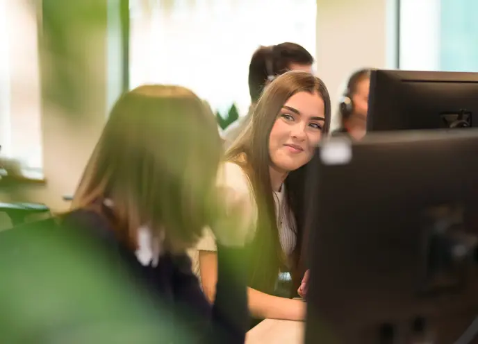 A group of women sitting at a desk with computers, one female smiling at the other.
